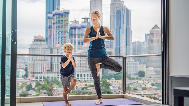 Mom and son are practicing yoga on the balcony in the background of a big city. Sports mom with kid doing morning work-out at home. Mum and child do the exercises together, healthy family lifestyle concept.