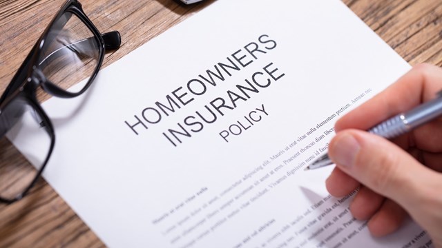 Overhead View Of A Person's Hand Filling Homeowners Insurance Policy Form Over Wooden Desk