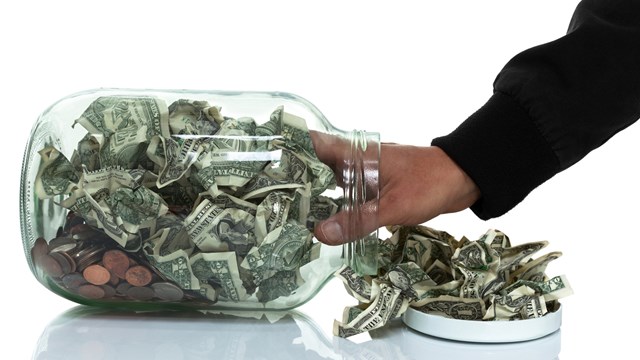 Glass jar full of money tipped over on its side spilling money with hand reaching in