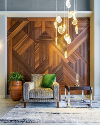 Interior of modern beige armchair, small wooden modern table, planter with green bushes, and contemporary tall glass chandelier, in a hall with decorated wood cladding wall, and white marble floor