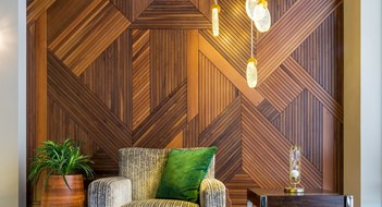 Grey is Gone in '24 Interior of modern beige armchair, small wooden modern table, planter with green bushes, and contemporary tall glass chandelier, in a hall with decorated wood cladding wall, and white marble floor