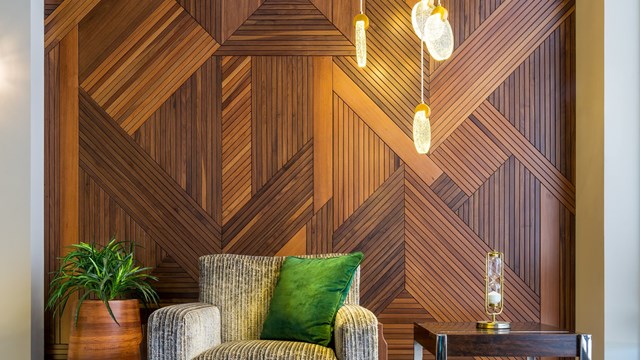 Interior of modern beige armchair, small wooden modern table, planter with green bushes, and contemporary tall glass chandelier, in a hall with decorated wood cladding wall, and white marble floor