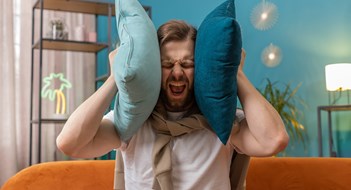 Q&A: Curbing the Complaints Repair work at neighbours. Irritated young man relaxing on couch cover ears with pillows annoyed by noisy neighbors suffer from headache wish silence. Thin walls at home flat without sound insulation