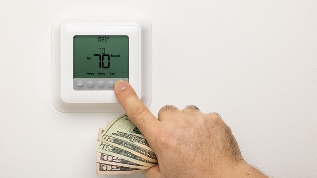 A hand holding money adjusting a thermostat on a wall.