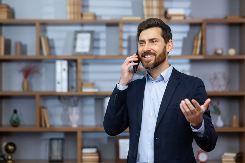 Mature successful boss happy with achievement talking on phone inside office, senior lawyer consulting clients on phone, businessman wearing glasses in business suit, man at workplace standing.