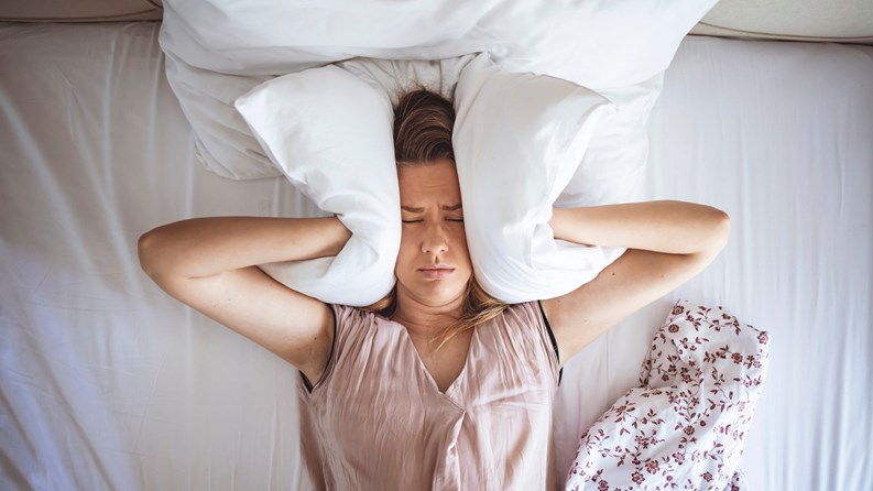 Young woman trying to sleep but disturbed by noisy neighbors and covering ears with pillows. I can's sleep with all that noise! Sleep problems. Angry woman disturbed with a noise trying to sleep